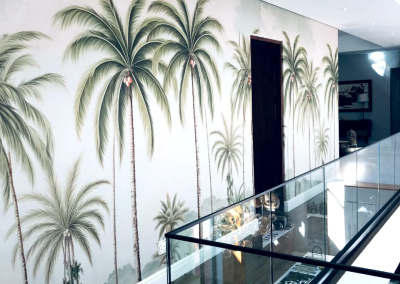 The tropical palm tree mural features lush green palm fronds with varying shades from light to dark green, set against a serene blue sky backdrop. The mural includes subtle hints of brown on the tree trunks and soft beige accents that mimic sunlight filtering through the leaves. This blend of natural elements and colors creates an inviting, tranquil atmosphere on the hallway wall.