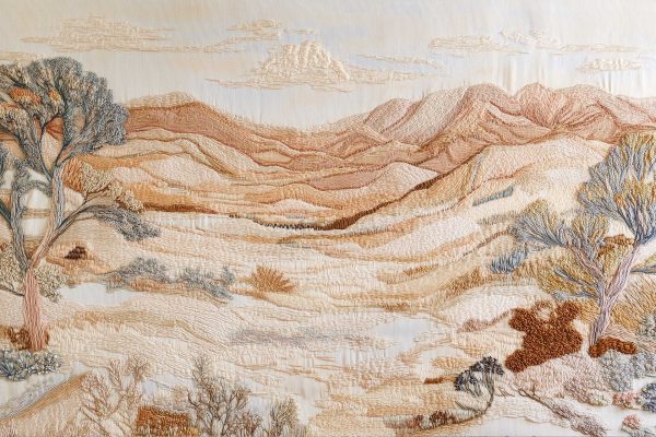 Wallpaper features an embroidered landscape with rolling hills and mountains in earthy tones. Includes two tall trees, low shrubs, and lightly embroidered clouds in the sky, conveying a serene desert ambiance. Colors: earthy browns, greens, soft blues.